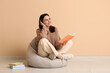 © Pixel-Shot - Young thoughtful woman sitting on pouf with book near beige wall