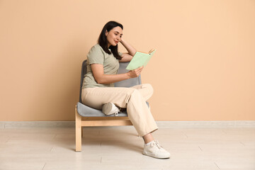 Canvas Print - Young woman sitting on armchair and reading book near beige wall