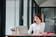 © Satori Studio - A young woman in a white shirt works on her laptop in a modern office setting with large windows and natural light.