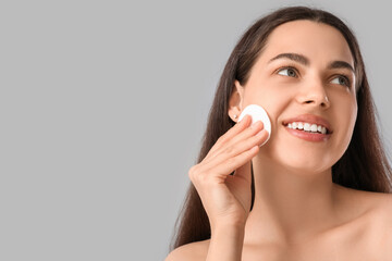 Wall Mural - Beautiful young happy woman with cotton pad removing makeup on grey background