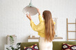 © Pixel-Shot - Young woman screwing light bulb into ceiling lamp at home, back view