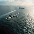 © Elena L - A drone soaring over open ocean waters, trailing a wake, with an aircraft carrier visible in the distance under a partly cloudy sky.