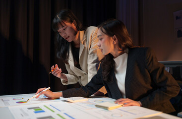 Wall Mural - Two businesswomen working overtime, having discussions, talking, brainstorming, pointing at graphs and charts to analyze online market data, doing calculations, accounting to plan company strategies.