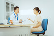 © Phushutter - A male Asian pediatrician in a white lab coat sits at his desk, warmly greeting a middle-aged pregnant woman for a prenatal consultation, offering care and support in a clinical setting.