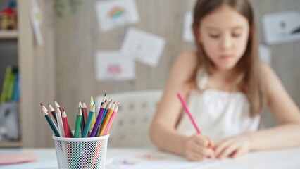 A close-up of colored pencils on the desk, with a creative young girl drawing in the background