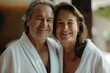© ChaoticMind - A senior couple wearing white bathrobes are smiling warmly at the camera, depicting a harmonious and loving relationship, as they pose together indoors in a soft light setting.