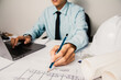 © PhotoJuthamat - architect meticulously reviews blueprints on a laptop, planning the design and construction of a new home, surrounded by sketches and tools in an office workspace.