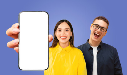 Fototapete - A woman holds a smartphone with a white blank screen in front of her, smiling at the camera. A man wearing glasses and a dark shirt stands behind her, smiling and looking at the camera.