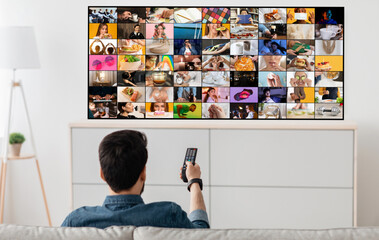 A man sits on a couch in a living room, watching television. The television screen shows multiple channels playing simultaneously, displaying a variety of content