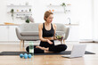 © sofiko14 - Woman in activewear sitting on yoga mat eating salad while using laptop. Exercise equipment and water bottle on floor. Home interior with cozy setting.