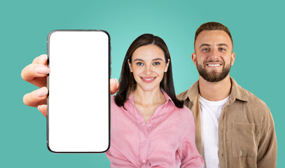 Fototapete - A couple, a woman in a pink shirt and a man in a tan corduroy jacket, stands in front of a teal background, smiling. The woman is holding a smartphone with a blank white screen, facing the camera.