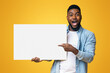 © Prostock-studio - Joyful black man pointing at blank white advertising board in his hands, yellow studio background with free space