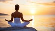 © VISUAL BACKGROUND - A woman sitting in a lotus position on a rock by the water at sunset