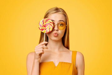 Wall Mural - Sweet Treat. Pretty young summer girl covering eye with big bright lollipop and looking at camera, yellow background with copy space