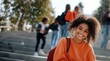 © David Giraud - Une jeune étudiante afro-américaine heureuse et souriante, assise dans les escaliers à l'université, portant une veste orange et un sac à dos.