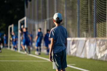 boy standing and looking at football players, blue football players uniform, football field, game, childhood, child, baby, kid,