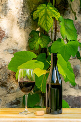 A bottle and a glass of red wine on a wooden table. Stone wall and the leaves of a vine in the background. Copyspace