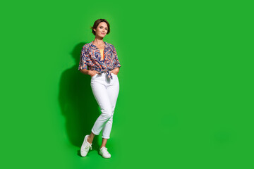 Poster - Full size photo of adorable pleasant girl wear print shirt standing near empty space palms in pockets isolated on green color background