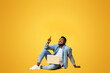 © Prostock-studio - Great offer. Black millennial guy sitting on floor with laptop and pointing upwards at copy space on yellow background