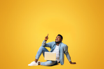 Wall Mural - Great offer. Black millennial guy sitting on floor with laptop and pointing upwards at copy space on yellow background