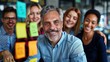 © Pinklife - A cheerful team of professionals brainstorming in an office, with a mature man at the front and sticky notes on the wall, indicating collaboration and creative thinking.