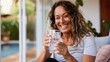 © Pinklife - A cheerful woman with waves in her hair is holding a glass of water, smiling happily while relaxing on an outdoor patio, bathed in sunlight and surrounded by nature.