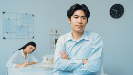 Wall Mural - Professional asian businessman crosses arms with confident while young beautiful caucasian colleague drawing a blueprint on meeting table with house model and blueprint at modern office. Immaculate.