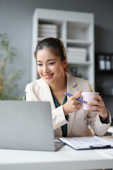 Asian businesswoman is smiling while drinking coffee and working on a laptop in her office