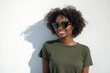 © GustavsMD - A cheerful Black woman wearing sunglasses and an olive green t-shirt smiles confidently while standing against a bright white wall, casting a soft shadow