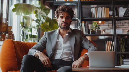 A man in a suit sits on an orange couch in front of a laptop