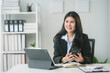 © Crystal - Asian businesswoman is holding a smartphone and smiling while sitting at her desk in a modern office