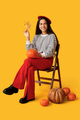 Wall Mural - Stylish young woman with fresh pumpkin and autumn leaf sitting on chair against yellow background