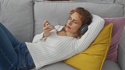 Wall Mural - A relaxed woman lounges on a couch at home with headphones and smartphone, embodying leisure in a casual setting.