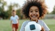 © Lens Legacy - A joyful child with curly hair is holding a soccer ball with a big smile on their face while in a park, presenting a scene filled with happiness, play, and outdoor activity.