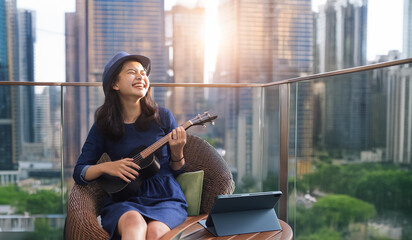 asian girl having fun play ukulele and sign a song on  from tablet on balcony high residence in city scape