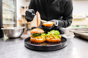 Wall Mural - A chef in a black uniform assembles a gourmet burger with fresh ingredients on a black plate in a professional kitchen. The close-up highlights culinary skills and attention to detail.