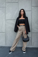 Wall Mural - Fashionable beauty woman with curly hair in trendy urban clothes with blazer, top, bell-bottom pants, shoes and bag stands near a gray wall in the city