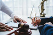 © amnaj - Lawyer holding pen is signing important legal contract with client in office with judge gavel and themis statue in background