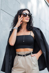 Wall Mural - Fashion beautiful young curly elegant woman with sunglasses in black trendy stylish clothes with blazer and top with pants sits near gray building in city