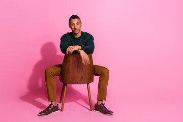 Full length photo of handsome good mood guy wear sweatshirt sitting armchair smiling emtpy space isolated pink color background