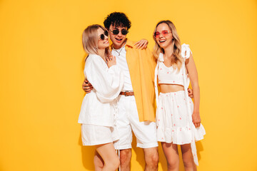 Group of young three stylish friends posing in studio. Fashion man and two cute female dressed in casual summer clothes. Smiling models having fun. Cheerful women and guy near yellow wall, sunglasses