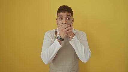 Canvas Print - Shocked young hispanic man wearing apron, covering mouth with hands over a mistake, standing isolated on yellow background - the secret shock