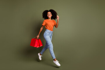 Poster - Full length photo of charming positive woman wear orange t-shirt walkign rising shoppers empty space isolated khaki color background