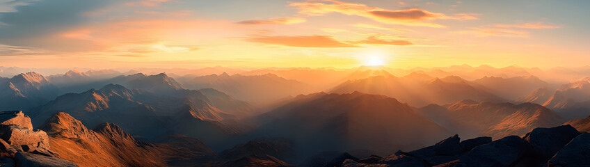 Wall Mural - A beautiful sunset over a mountain range with a clear blue sky. The sun is setting and the sky is filled with clouds. The mountains are covered in rocks and the landscape is breathtaking