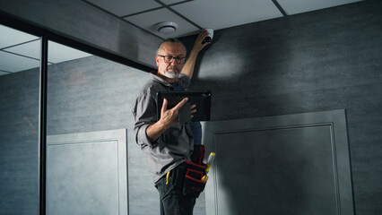 male installer adjusts the angle of cctv camera in office, sets up it using digital tablet computer 