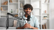 © YURII MASLAK - Microphone, podcast and Indian man speaker in his living room. Creative streamer, internet influencer or online content creator live streaming for audience. Camera view.
