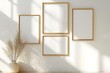 © Kakabe - Four golden picture frames of various sizes hang on a sunlit white wall, with a vase containing dry reeds placed below, Ideal for gallery design, home decor, or art presentation visuals,