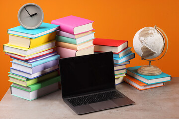 Wall Mural - Stacks of colorful books, globe, clock and laptop on wooden table against orange background