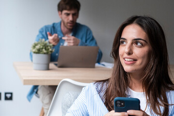 Young and modern woman in casual clothes smiling and using her smartphone to scroll social media or read content while working in the office, with her colleague engaged in a video call on a laptop 