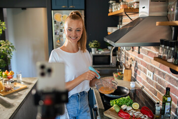 Smiling young woman cooking and filming with smartphone in modern kitchen